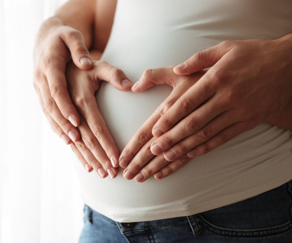 Close up portrait of man's and woman's hands making heart gesture over pregnant belly indoors