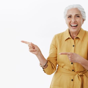 Portrait of delighted healthy and active pretty senior female in yellow trendy coat and bracelet laughing out loud from amusement standing entertained and charistmatic pointing left with forefingers.